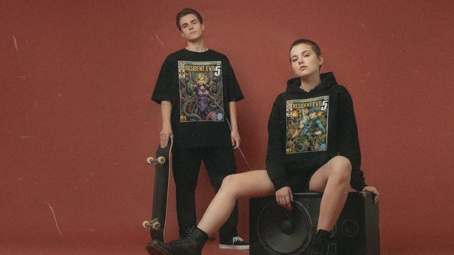Two models wearing SukiPrime streetwear against a red background. The male model wears an oversized black t-shirt with a Resident Evil retro comic cover art print. The female model wears a black hoodie with matching gaming comic art.